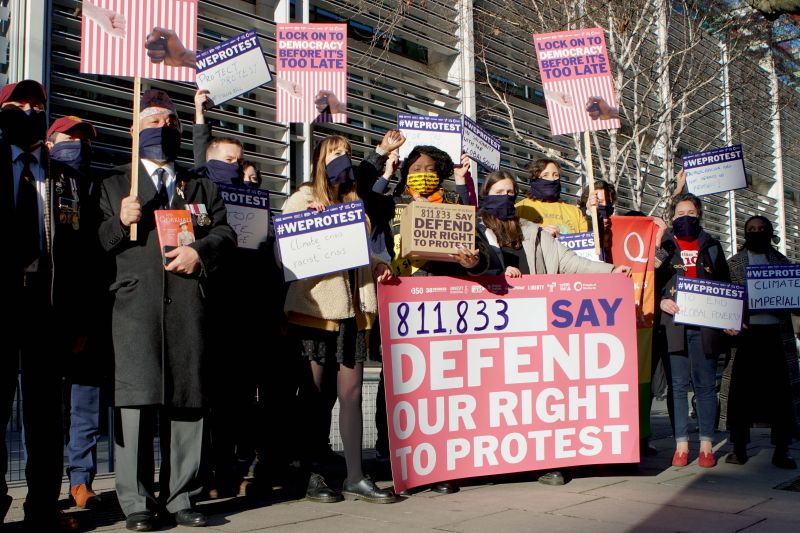 A group of people, different ages and ethnicities, wearing face masks and holding placards. The main placard reads "811,833 say defend our right to protest' and others are holding smaller placards with reasons why they are protesting which aren't legible.