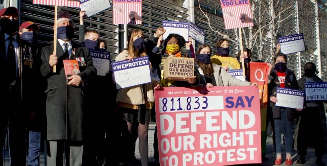 A group of people, different ages and ethnicities, wearing face masks and holding placards. The main placard reads "811,833 say defend our right to protest' and others are holding smaller placards with reasons why they are protesting which aren't legible. 