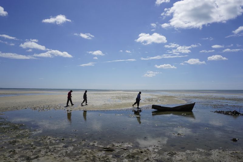 A beach photo showing off the sky, with 3 people in the distance and a boat which is not in the water, but on the dirty looking sand