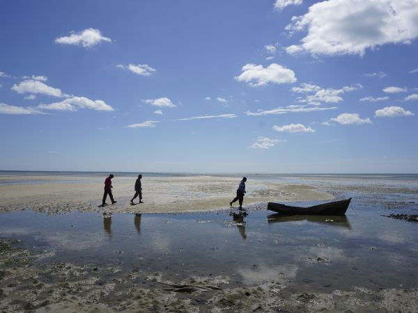 A beach photo showing off the sky, with 3 people in the distance and a boat which is not in the water, but on the dirty looking sand