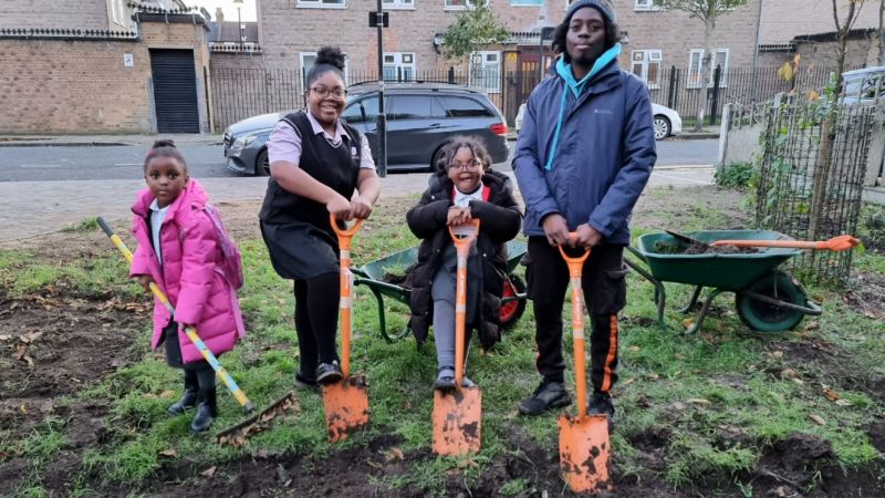 Four children with orange spades digging into grass with wheelbarrows behind them