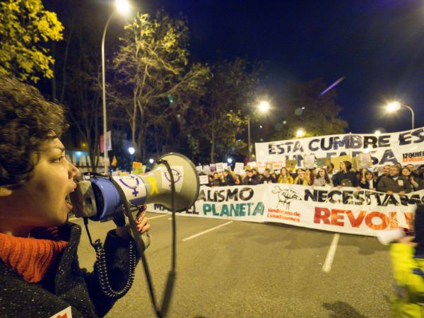 A young woman holding a megaphone, mid-speaking, facing an ongoing rally holding protest signs.