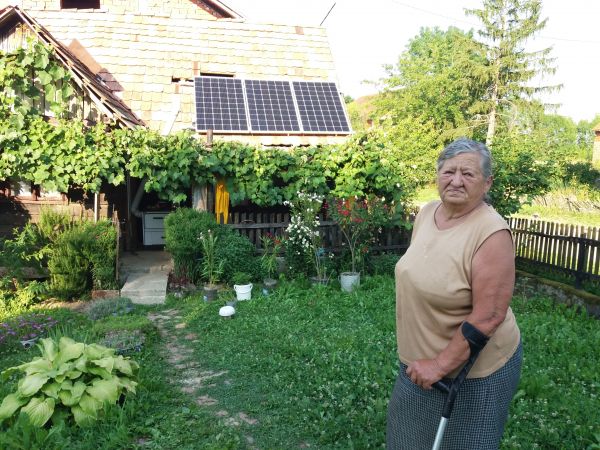An elderly woman in her green garden, she has a walking aid. She is looking at the camera, and in the back is her home with solar panels.