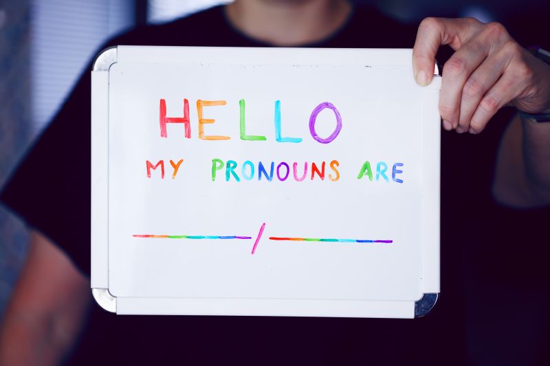Someone is holding up a beautiful, rainbow-lettered placard which reads "Hello my pronouns are" with space to fill in your gender pronouns.