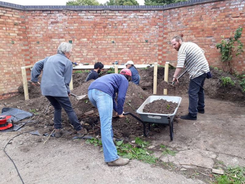 A group of 5 people filling a wheelbarrow with soil