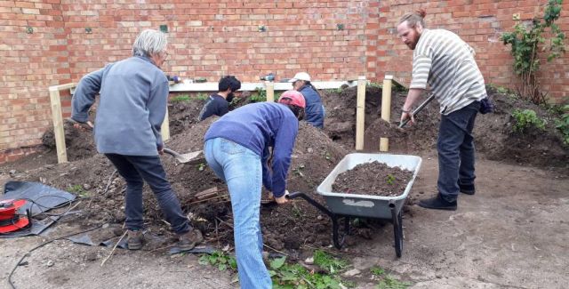 A group of 5 people filling a wheelbarrow with soil