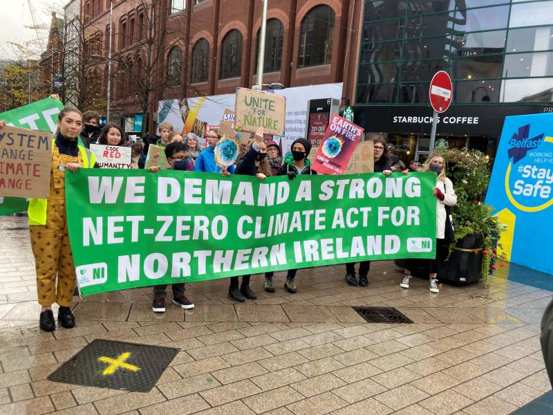 Protesters outside in Belfast City Centre holding a sign up saying 'We demand a strong net-zero climate act for Northern Ireland'