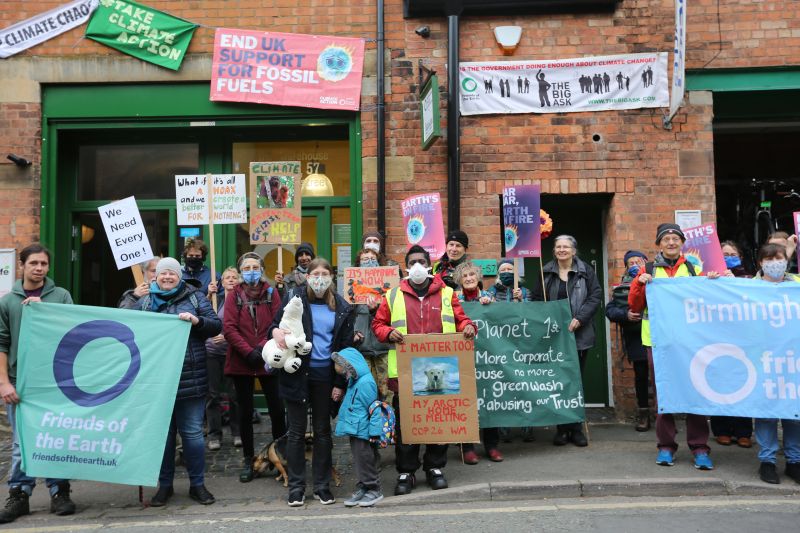 A group of climate protesters of different ages and ethnicities standing outside a building with friends of the earth and climate change banners