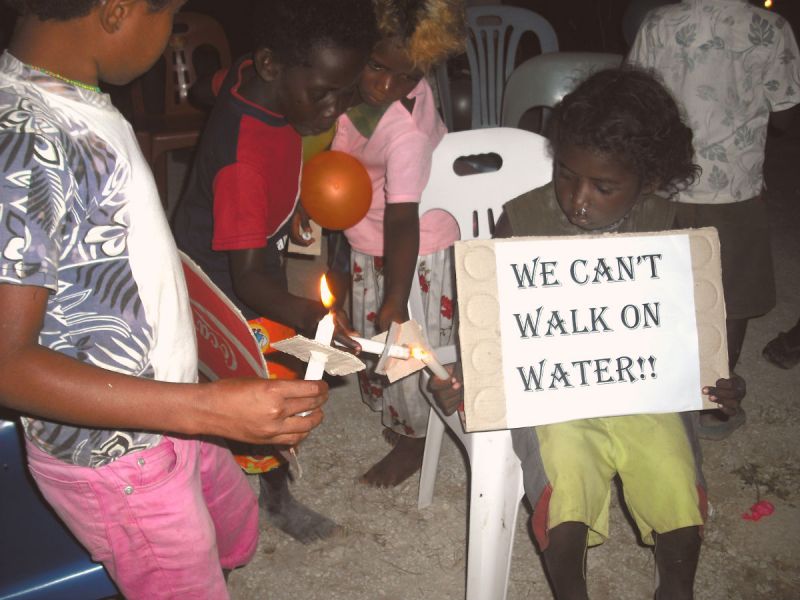 Children lighting candles in Papa New Guinea. One holds a sign saying "We can't walk on water!!"