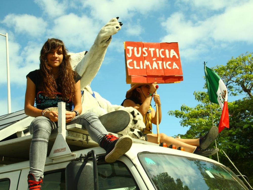 Two young women sitting on top of a van holding a placard which reads justicia climática