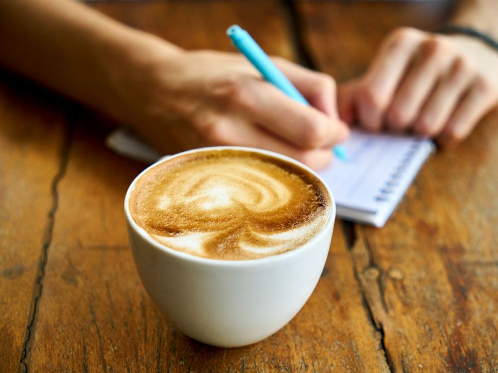 A close up of a man writing in a notepad with a cup of cappuccino