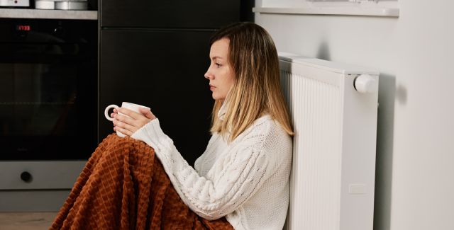 Worried-looking woman sat by radiator, wrapped in a blanket and drinking tea