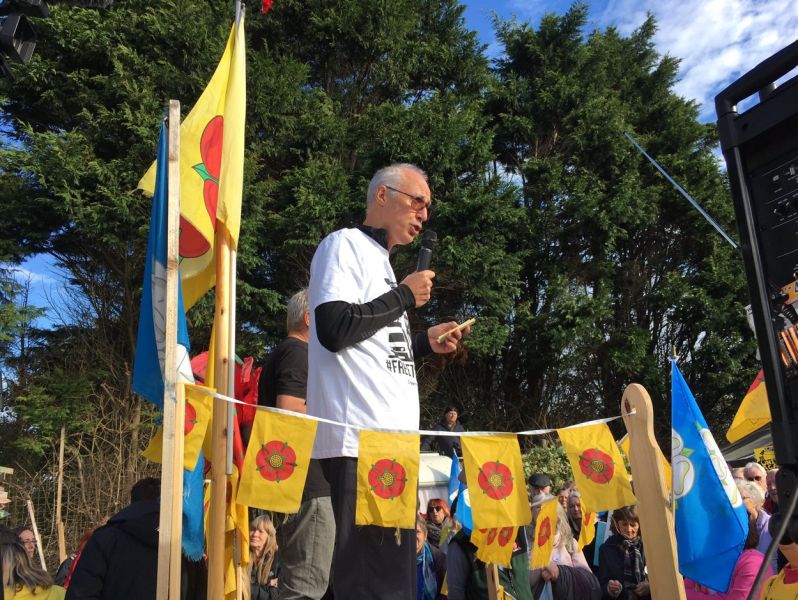 A man holding a mic speaking at a rally, there are flags with flowers present, and an audience