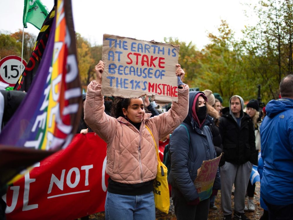 A shot of a protest with focus on a young woman holding a banner which reads "the earth is at stake because of the 1%'s mistake"J