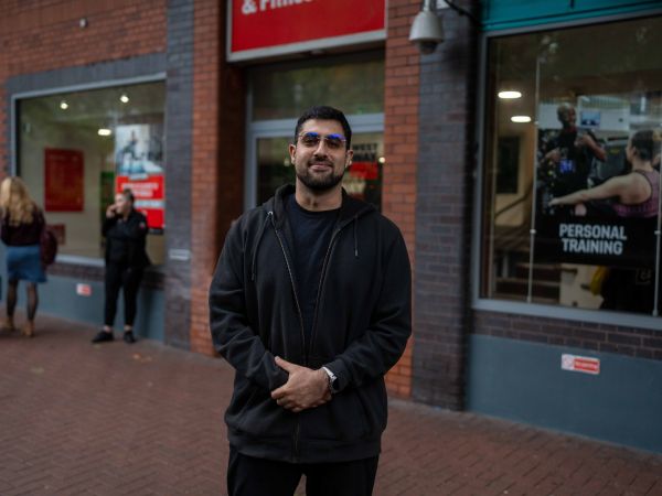 A man dressed in black, looking relaxed facing the camera and smiling. He's stood outside a gym.