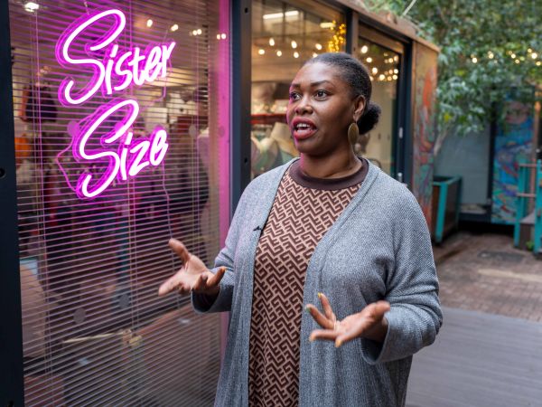 A woman stood outside her business "Sister Size" - her business sign is lit of in neon and she's looks like she's mid-speech and has expressive hands