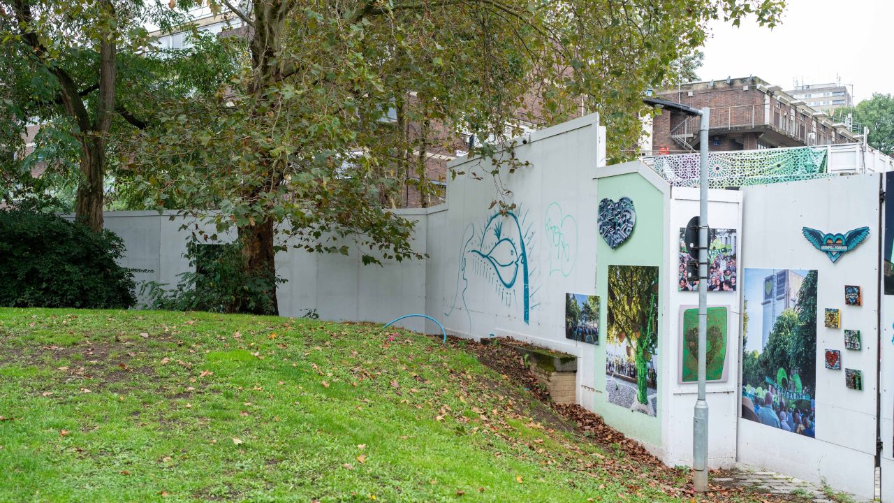 An park in an urban area. There's a wall which has images on it remembering the Grenfell tragedy, as well as drawn illustrations of nature. There's a couple of trees too.