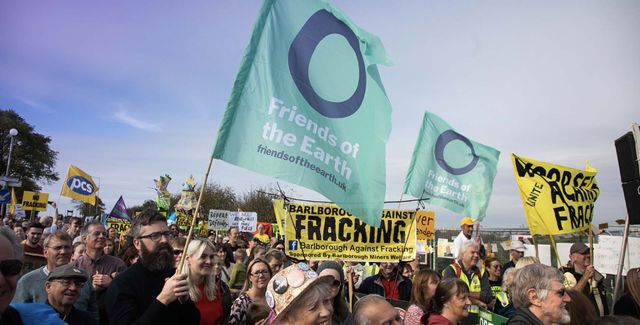 A large protest crowd, people of all ages are looking joyful and are carrying friends of the earth flag and anti-fracking banners.