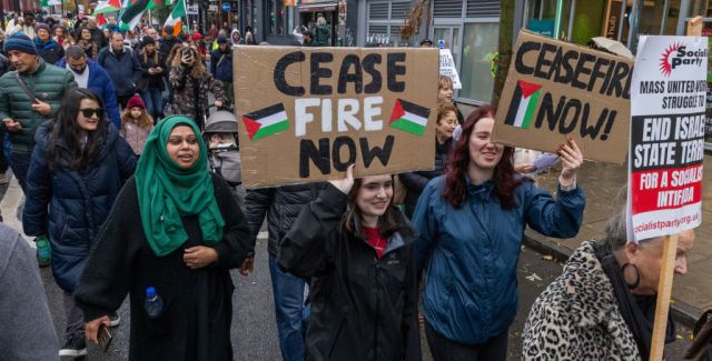 A crowd marches along a London street, 2 women in the foreground hold placards with "Ceasefire now" and the Palestinian flag painted on them