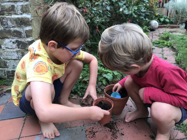 Two little boys are planting a seeded paper shape into plant pots. The older boy has brown hair and wears a yellow tshirt and blue glasses. The younger has a long-sleeved red top and sandy blonde hair.