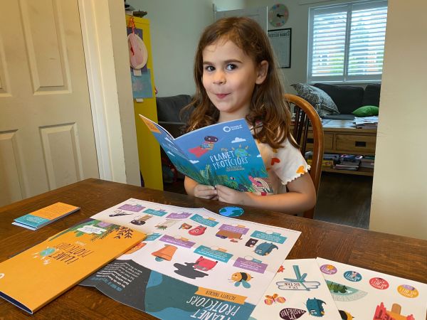 A little girl with brown hair, brown eyes and white skin sits at a wooden table. She holds her activity book up, and looks cheekily at the camera. The rest of the pack is on the table in front of her. She is wearing a white top with little yellow and red hearts on it.