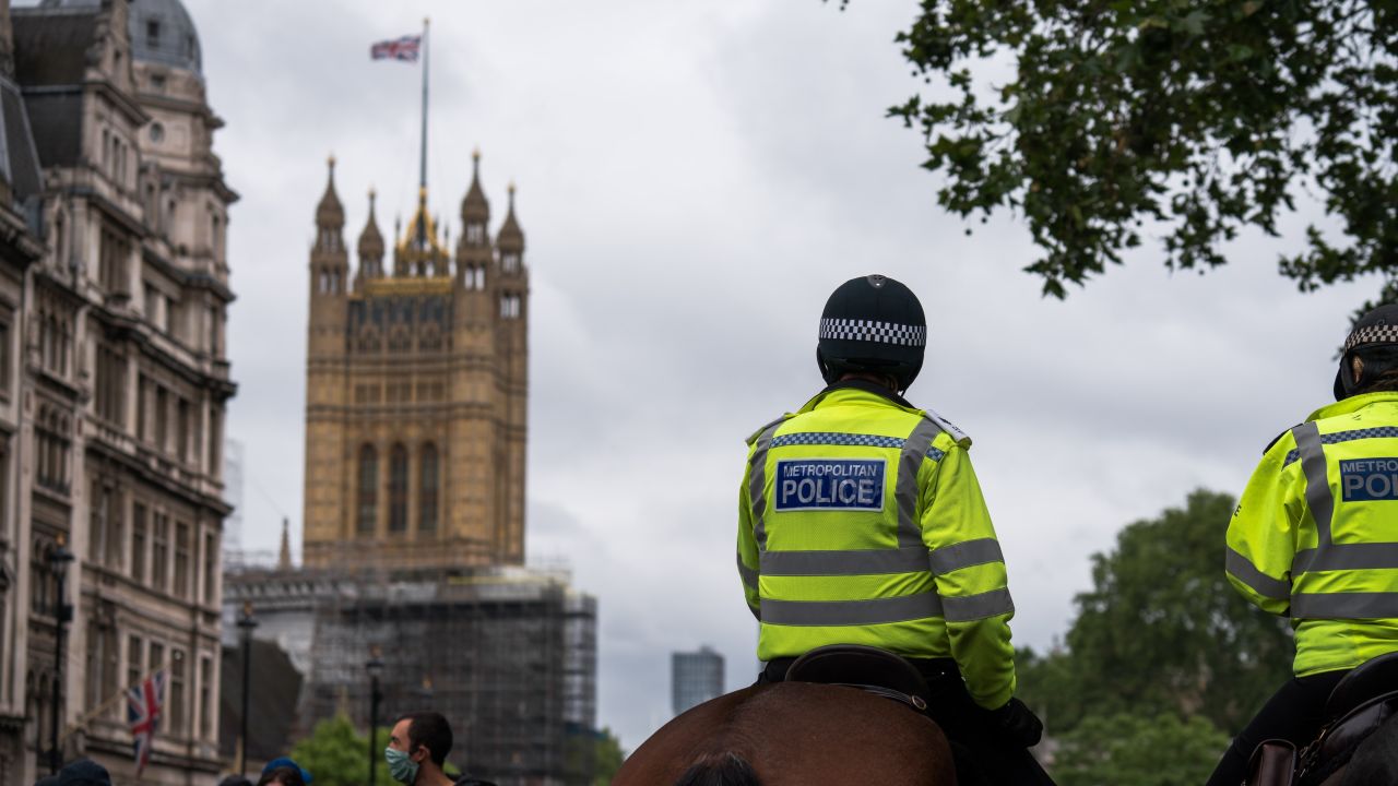 The backs of two police men on horses, with London scenery in the background where we can see a waving British flag on an old building and people in face masks.