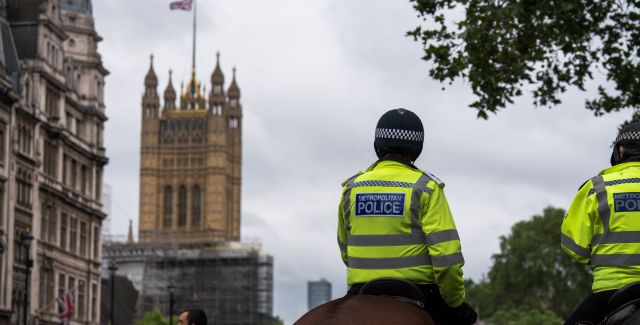 The backs of two police men on horses, with London scenery in the background where we can see a waving British flag on an old building and people in face masks.