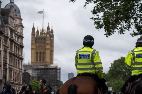 The backs of two police men on horses, with London scenery in the background where we can see a waving British flag on an old building and people in face masks.