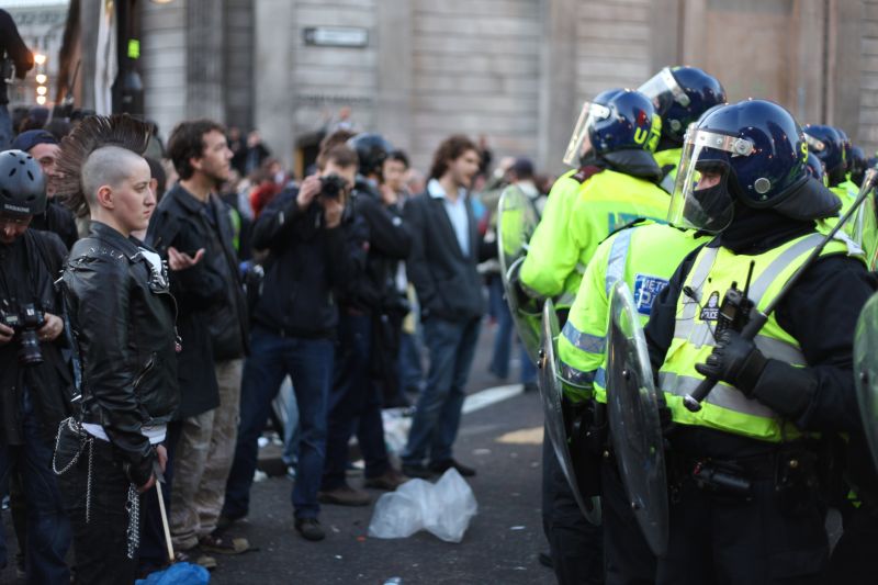 A protest stand off - a person is looking directly at a line of police officers with batons and protective gear