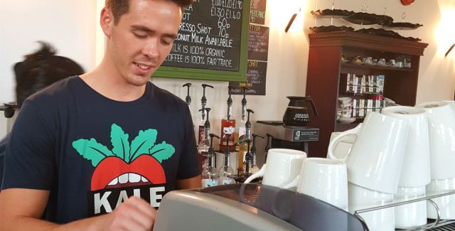A member of staff wearing a Kale Yeah! t-shirt serving students at a coffee bar counter at Portsmouth University