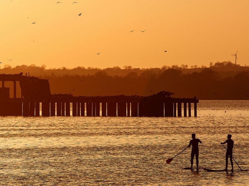 Two silhouetted figures at sunset doing stand-up paddling in front of a derelict torpedo-testing platform in Lough Neagh