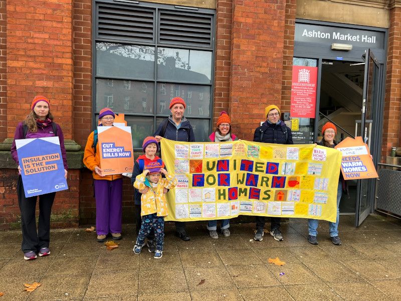 A group of people of various ages, including children, outside their MPs office with placards demanding warm homes and lower energy bills