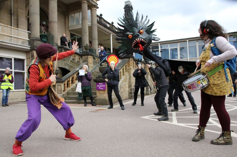A group of people performing a stunt outside a town hall, including a large oil monster and drumming