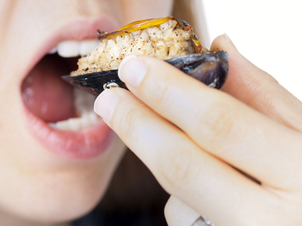 Close-up of a woman's hand and mouth as she eats a dressed mussel from its shell