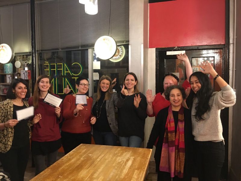 A group of women stand round a table smiling and with their hands raised in celebration
