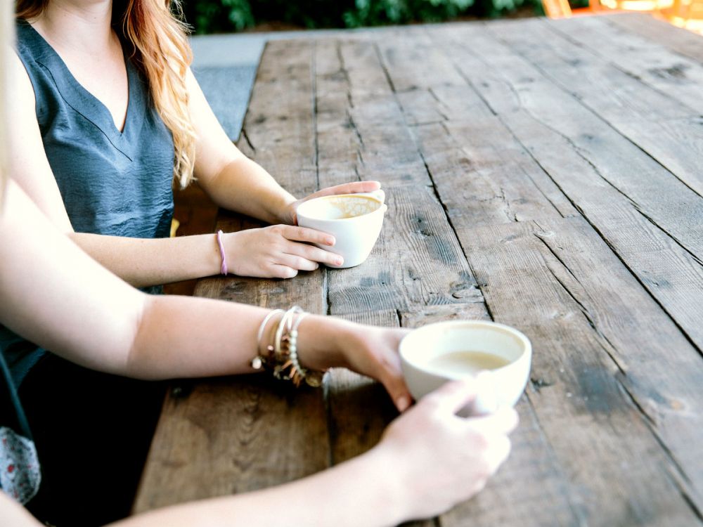 Two women at a cafe drinking coffee at a long wooden table.