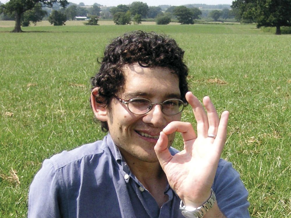 Phil Michaels in a green field with trees in the background, smiling and making the Friends of the Earth logo with his hand