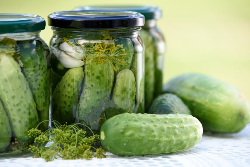 Close-up of 3 jars of homemade plastic-free pickled gherkins with 3 fresh gherkins next to them.