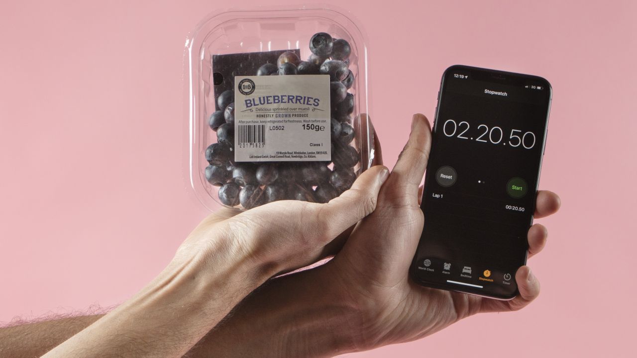 One hand holding a smartphone showing a timer screen and the other hand holding a plastic tub of blueberries