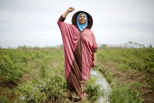A woman in a hijab and jilbaab standing on land with her fist in the air, and a smile on her face.