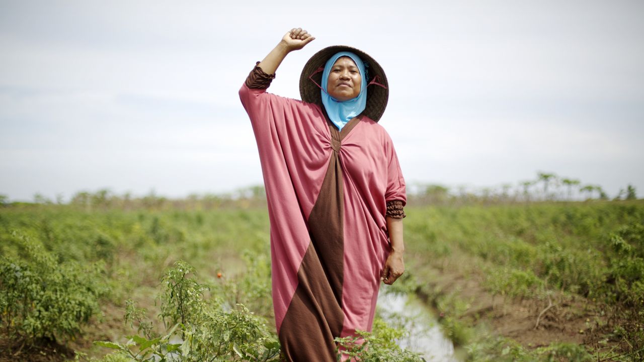 A woman in a hijab and jilbaab standing on land with her fist in the air, and a smile on her face.