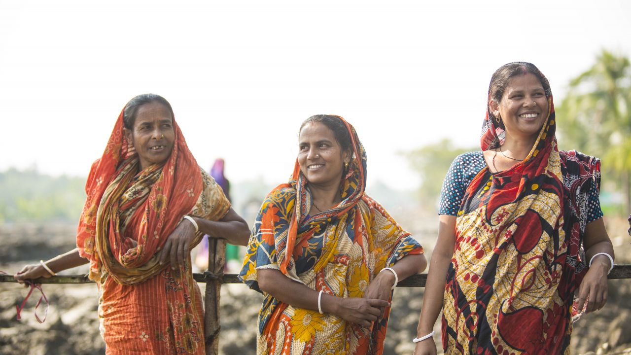 Three women in bright saris standing against a fence with big smiles