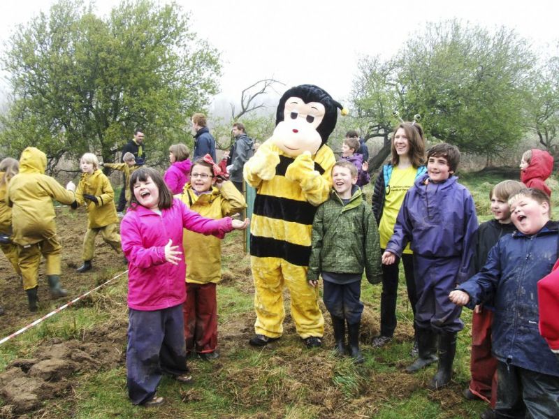 Person in bee costume and lots of children in field with trees in background