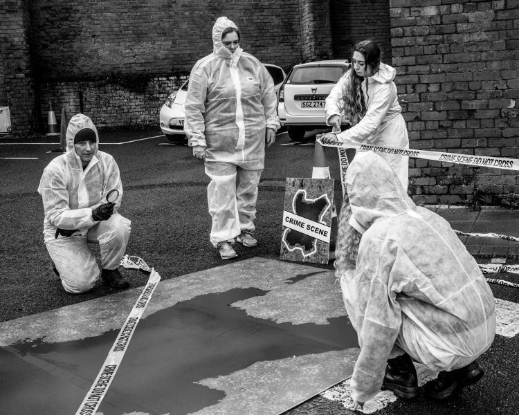 A staged crime scene. People are wearing protective white suits and are laying out criminal tape. Black and white image, very moody and dark.