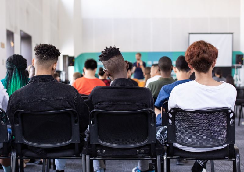 Photo taken from behind. Teenage students sitting on chairs facing the teacher.