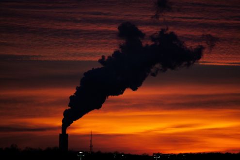 A photo taken at dusk showing a silhouette of the top of a factory emitting a lot of thick smoke
