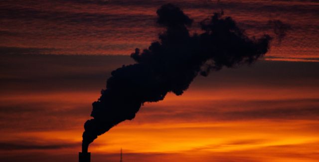 A photo taken at dusk showing a silhouette of the top of a factory emitting a lot of thick smoke