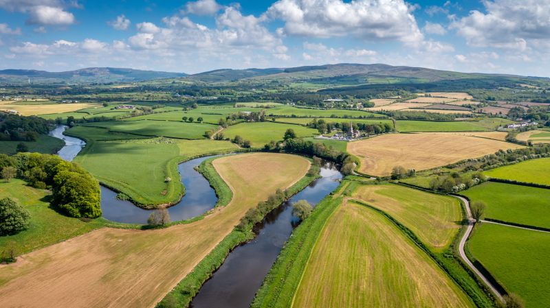 A photo of a scenic landscape which looks still and undisturbed. Gorgeous well kempt fields, and streams of water. In the distance, there's mountains amongst a peaceful blue sky.