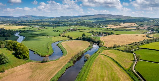 A photo of a scenic landscape which looks still and undisturbed. Gorgeous well kempt fields, and streams of water. In the distance, there's mountains amongst a peaceful blue sky.
