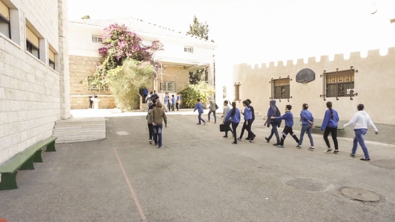 A school playground, with young children in blue uniform walking straight in a line. Teachers are present. The photo is taken from the back.